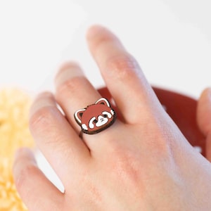 May include: A wooden ring featuring a red panda face with white accents. The ring is worn on a person's finger.