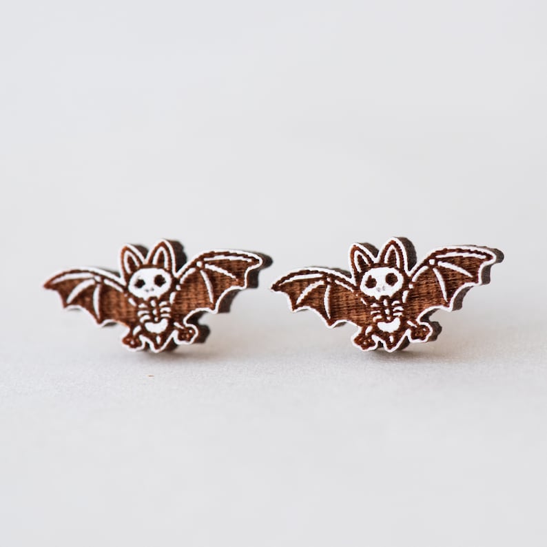 Peut inclure: Une paire de boucles d'oreilles en bois brun en forme de chauves-souris avec des d&eacute;tails peints en blanc. Les chauves-souris ont un motif de cr&acirc;ne et d'os crois&eacute;s.