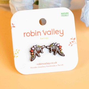 May include: A pair of wooden dinosaur skeleton earrings with rainbow colored accents. The earrings are on a white card with the text "robin valley" and "Earrings" printed on it. The card also says "Nature Friendly" and "Wooden Jewellery Handmade in the UK".