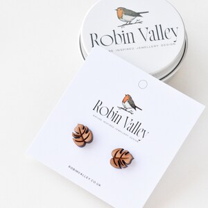 May include: A pair of wooden earrings shaped like monstera leaves. The earrings are brown and have a natural wood finish. The earrings are packaged in a small tin with a robin illustration and the text "Robin Valley" on it.