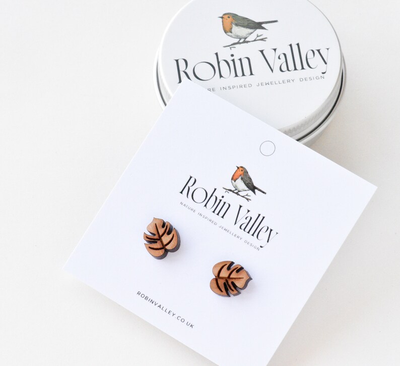 May include: A pair of wooden earrings shaped like monstera leaves. The earrings are brown and have a natural wood finish. The earrings are packaged in a small tin with a robin illustration and the text "Robin Valley" on it.