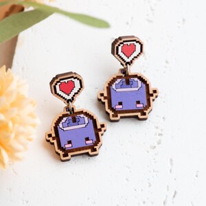 May include: A pair of pixelated wooden earrings. Each earring features a purple robot-like figure with a red heart above it. The earrings are made of wood with a pixelated design and a brown border.