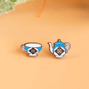 Op de afbeelding: Een paar blauwe en witte keramische oorbellen in de vorm van een theekopje en een theepot. Het theekopje en de theepot hebben een gouden en zwart geometrisch ontwerp.