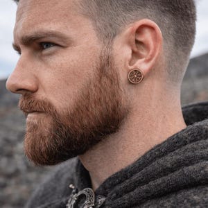 May include: A circular wooden stud earring featuring a detailed laser-engraved Vegvisir compass symbol. The earring has a natural light brown wood grain finish and is worn in the earlobe, showcasing a precise, intricate design on its flat surface.