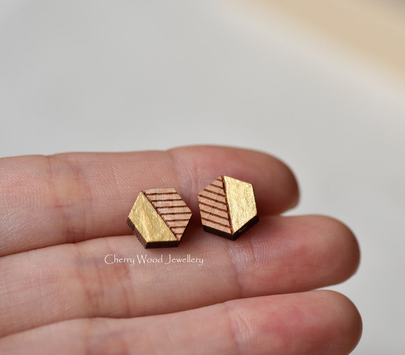 May include: A pair of hexagonal wooden earrings with a gold geometric design. The earrings are a light brown color with gold accents.