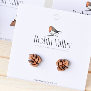 May include: A pair of wooden earrings featuring a brown monstera leaf design. The earrings are displayed on a white card with the text "Robin Valley Nature Inspired Jewellery Design" and "robinvalley.co.uk".