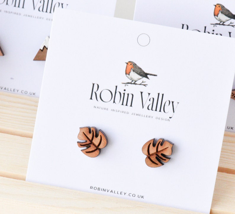 May include: A pair of wooden earrings featuring a brown monstera leaf design. The earrings are displayed on a white card with the text "Robin Valley Nature Inspired Jewellery Design" and "robinvalley.co.uk".