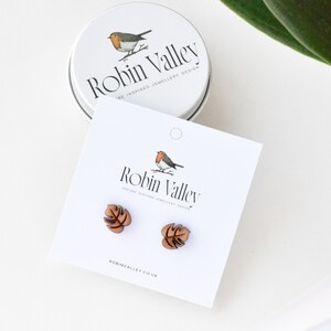 May include: A pair of wooden earrings shaped like monstera leaves. The earrings are brown and have a natural wood finish. The earrings are displayed on a white card with the text "Robin Valley" and "Nature Inspired Jewellery Design".