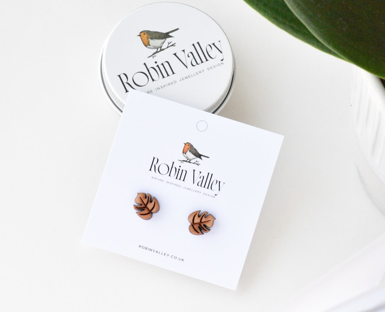 May include: A pair of wooden earrings shaped like monstera leaves. The earrings are brown and have a natural wood finish. The earrings are displayed on a white card with the text "Robin Valley" and "Nature Inspired Jewellery Design".