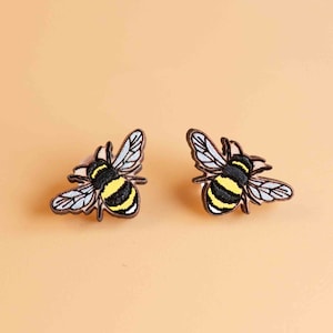 Peut inclure: Une paire de boucles d'oreilles en bois en forme d'abeilles. Les abeilles sont peintes avec des rayures noires et jaunes et ont des ailes brunes.