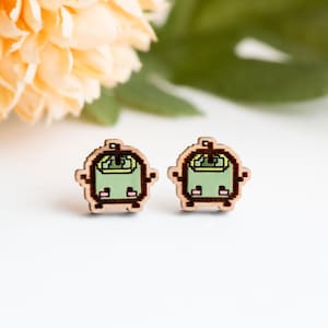 May include: A pair of wooden stud earrings with a pixelated green and brown design. The earrings have a light brown border and a green interior, with small pink details. The earrings are designed to look like a pixelated robot.