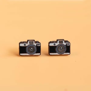 May include: A pair of miniature earrings shaped like cameras. They are black with white and grey detailing, including a lens and buttons. The background is a pale orange colour.