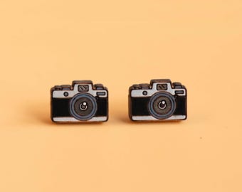 Op de afbeelding: Twee kleine, zwart-witte oorbellen in de vorm van een camera. De oorbellen hebben een gedetailleerd ontwerp, inclusief een lens, knoppen en een zoeker. De achtergrond is effen lichtoranje.