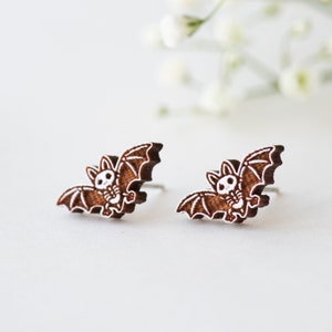 Peut inclure: Une paire de boucles d'oreilles en bois brun en forme de chauves-souris avec des d&eacute;tails peints en blanc. Les chauves-souris ont un aspect mignon et cartoon.