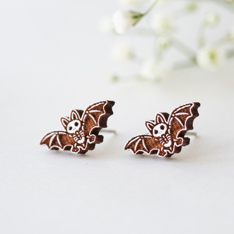 Peut inclure: Une paire de boucles d'oreilles en bois brun en forme de chauves-souris avec des d&eacute;tails peints en blanc. Les chauves-souris ont un aspect mignon et cartoon.