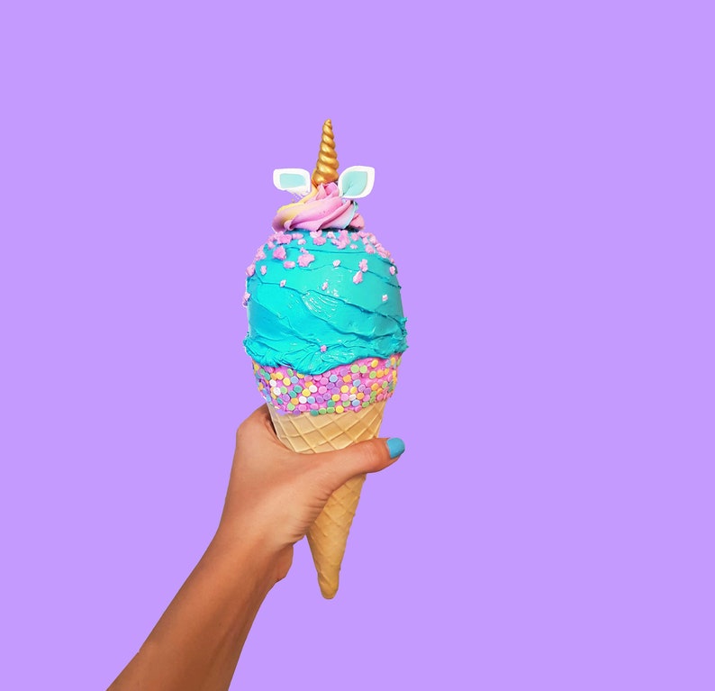 May include: A hand holds a blue and pink unicorn ice cream cone with sprinkles. The cone is on a purple background.