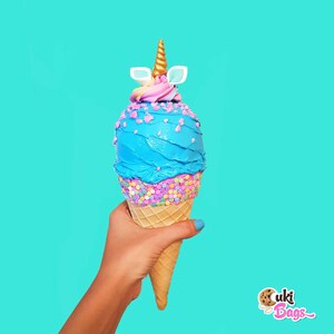 May include: A blue and pink unicorn ice cream cone with sprinkles. The cone is held by a hand.