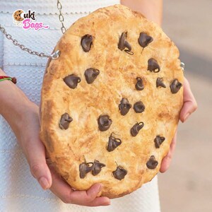 May include: A large, round, chocolate chip cookie purse with a brown chain strap. The purse is made to look like a real cookie with chocolate chips.