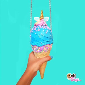 May include: A blue and pink ice cream cone shaped purse with a gold unicorn horn and white ears. The purse is hanging from a silver chain and is being held by a hand. The background is a bright turquoise color. The text "Cuki Bags" is visible in the bottom right corner of the image.