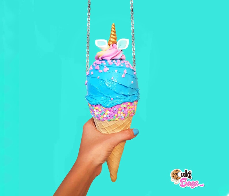 May include: A blue and pink ice cream cone shaped purse with a gold unicorn horn and white ears. The purse is hanging from a silver chain and is being held by a hand. The background is a bright turquoise color. The text "Cuki Bags" is visible in the bottom right corner of the image.