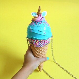 May include: A blue and pink ice cream cone shaped purse with a unicorn horn and ears on top. The purse has a silver chain strap.