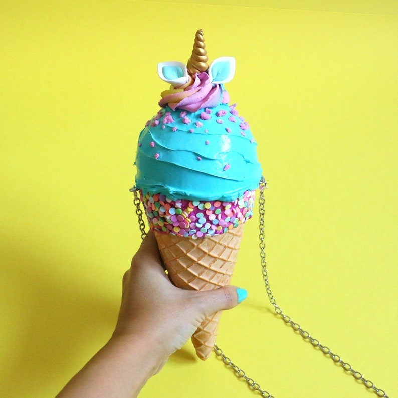 May include: A blue and pink ice cream cone shaped purse with a unicorn horn and ears on top. The purse has a silver chain strap.
