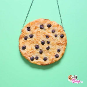 May include: A chocolate chip cookie necklace with a silver chain. The cookie is brown and has chocolate chips on it. The cookie is hanging from the chain.