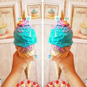 May include: Two turquoise blue ice cream cones with pink and white frosting and sprinkles. The cones have a unicorn horn and ears on top. The cones are being held by a hand.
