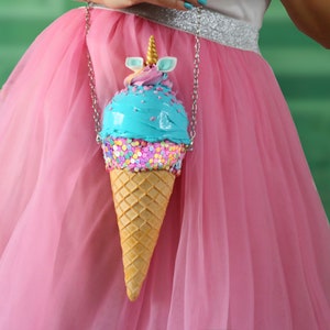 May include: A blue and pink ice cream cone shaped purse with a unicorn horn on top. The purse has sprinkles and a chain strap.
