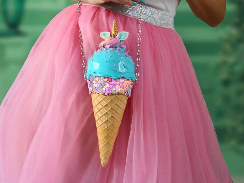 May include: A blue and pink ice cream cone shaped purse with a unicorn horn on top. The purse has sprinkles and a chain strap.