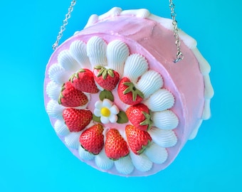 Puede incluir: Un bolso de pastel rosa con glaseado blanco y fresas rojas. El bolso tiene una correa de cadena plateada.