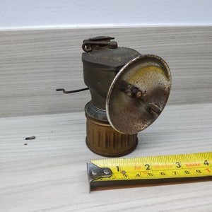 May include: A vintage brass miner's lamp with a round reflector and a handle. The lamp is sitting on a wooden surface next to a yellow measuring tape.