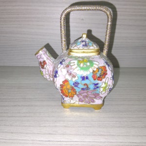 Puede incluir: Una tetera pequeña, blanca con un diseño floral. La tetera tiene un asa dorada y una tapa dorada. La tetera está decorada con flores y hojas de colores.