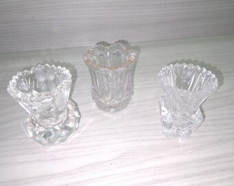 May include: Three clear glass bud vases with intricate cut designs. The vases are all different shapes and sizes, and they are all sitting on a wooden surface.