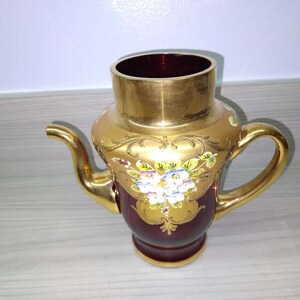 Puede incluir: Una tetera de vidrio roja con detalles dorados y diseños florales. La tetera tiene un asa y un pico.
