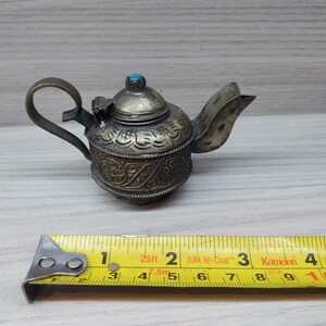 May include: A small, silver teapot with intricate floral designs and a turquoise stone on the lid. The teapot is about 2 inches tall and is sitting on a wooden surface.