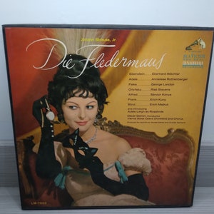 Puede incluir: Una portada de álbum de vinilo vintage con una mujer con un vestido negro y una máscara de carnaval. El álbum es para Die Fledermaus de Johann Strauss Jr., una opereta popular. La portada está en un esquema de colores naranja, rojo y amarillo vibrantes.