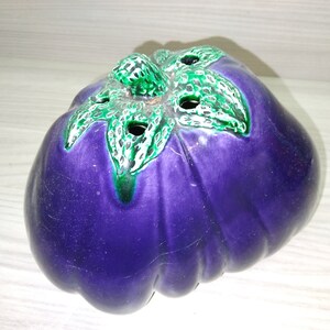 Könnte beinhalten: Eine violette, keramik-aubergine-förmige Vase mit grün-weißen bemalten Blättern auf der Oberseite. Die Vase hat Löcher oben für Blumen.