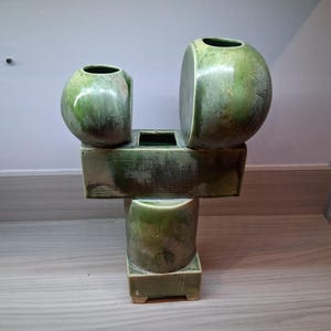 Puede incluir: Un jarrón de cerámica verde con un diseño abstracto único. El jarrón presenta tres secciones redondeadas, dos en la parte superior y una en la base, conectadas por bloques rectangulares. El jarrón tiene un acabado brillante y es adecuado para la decoración del hogar.