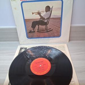 Puede incluir: Un álbum de vinilo vintage titulado "Louis Armstrong's Greatest Hits". La portada del álbum muestra a una persona tocando la trompeta. El disco es negro con una etiqueta roja y naranja. El álbum está en una funda blanca.