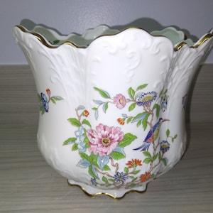 May include: White porcelain planter with a gold rim and floral design. The planter features a bird with blue, green, and yellow feathers perched on a branch with pink, blue, and yellow flowers.