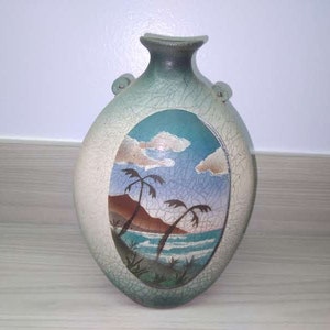 May include: A ceramic vase with a crackled glaze and a painted scene of palm trees and a beach. The vase is oval shaped with a narrow neck and two handles.