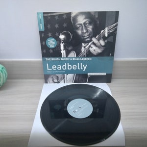 May include: A black vinyl record with a white label. The record is next to a book titled "The Rough Guide to Blues Legends: Leadbelly: Reissues and Remastered". The book cover features a black and white photo of a man playing a guitar.