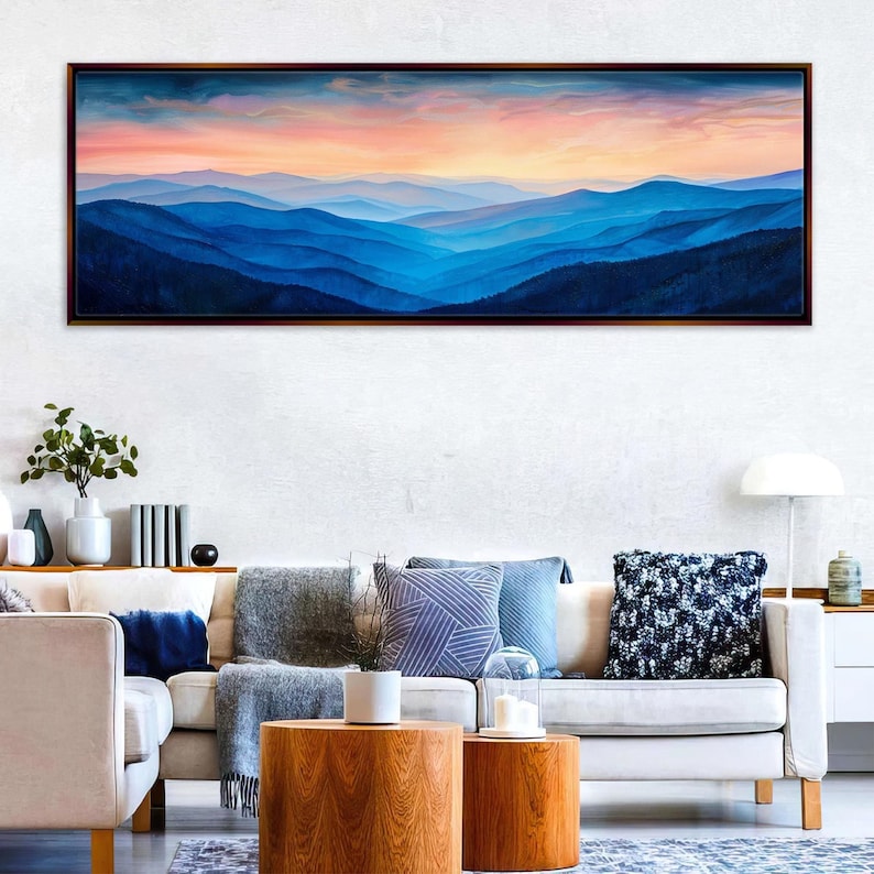 a large, framed painting of a mountain landscape with a colorful sunset over the mountains. The painting is hanging on a wall above a white couch with various throw pillows. The room has a modern and minimalist design, with a wooden coffee table in the center and a lamp on a side table.