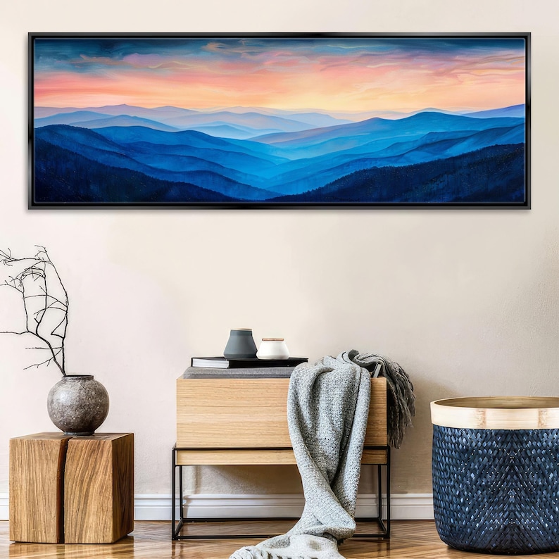 a large painting of a mountain landscape with a colorful sunset sky above. The painting is hanging on a wall above a wooden table with a vase and a book.