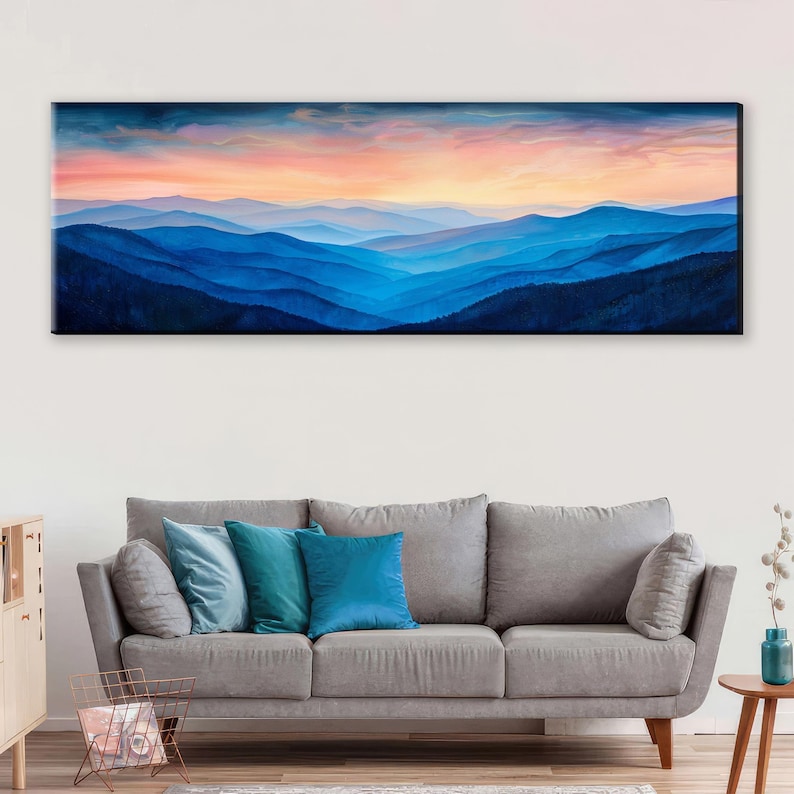 a large, long painting of a mountain range with a colorful sunset sky above it. The painting is hanging on a wall above a gray sofa with blue and teal throw pillows.