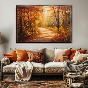 Könnte beinhalten: Ein gerahmtes Landschaftsgemälde zeigt eine Herbstwaldszene mit einem gewundenen Pfad. Das Kunstwerk weist warme Farben von Orange, Gelb und Braun auf, wobei das Sonnenlicht durch die Bäume scheint. Das Gemälde wird über einem Sofa mit orangefarbenen und beigen Kissen ausgestellt.