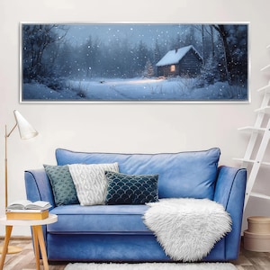 May include: A framed winter landscape painting depicts a snow-covered forest with a cozy cabin. The cabin's windows glow warmly against the backdrop of falling snow. The painting is displayed above a blue sofa with decorative pillows and a white fluffy throw.