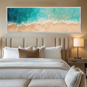 May include: A rectangular framed canvas art piece depicting an aerial view of a beach. The artwork features turquoise ocean waves meeting a sandy shore. The painting is framed in a white border and hangs above a bed with white linens and decorative pillows.