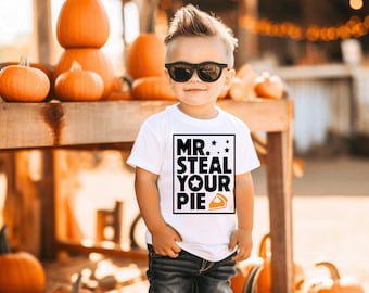 May include: A child wearing sunglasses and a white t-shirt printed with "MR. STEAL YOUR PIE". The child is also wearing distressed black jeans and white high-top trainers. The background includes pumpkins, suggesting an autumn theme.